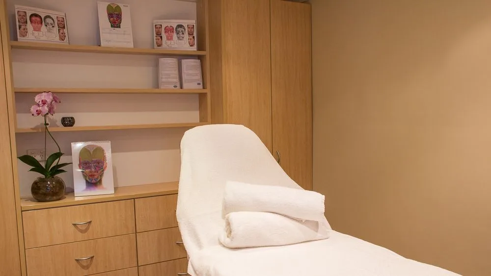 Treatment room: white-draped treatment bed, pale timber joinery, printed facial anatomy charts and a standing face-map flip chart, orchid