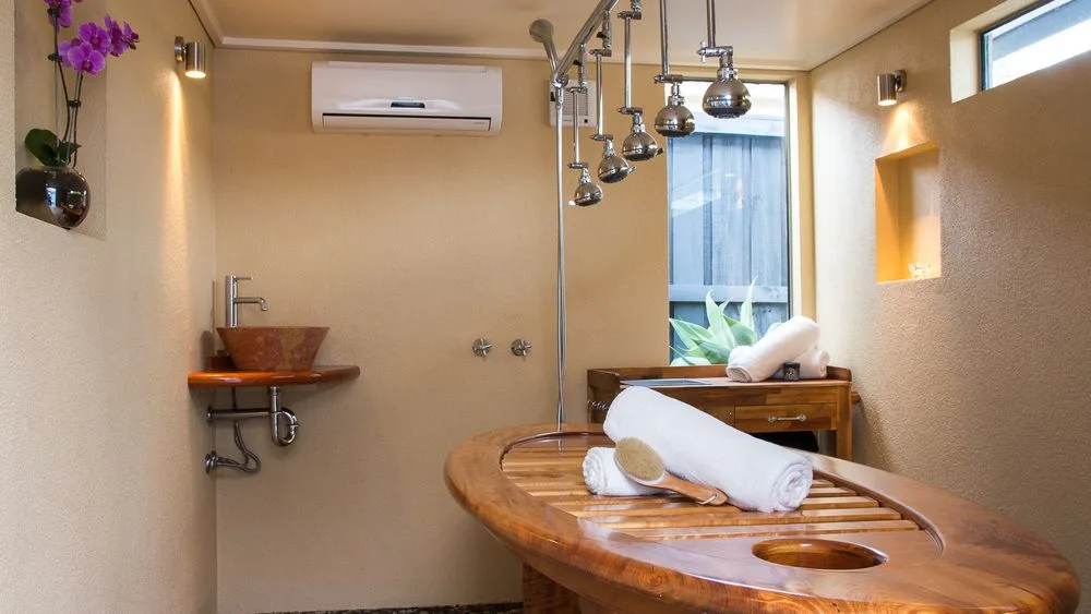 Wet room: seven-head Vichy shower over a carved timber table set with rolled white towels, sandstone-textured walls, orchid in a chrome vase