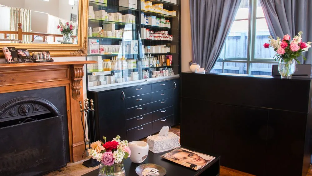 The clinic's front retail room: cast-iron fireplace with gilt overmantel mirror, floor-to-ceiling backlit skincare cabinet, roses on the low table