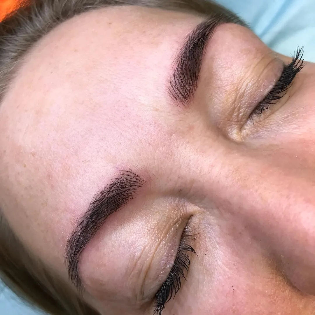 Finished feather-stroke brow tattooing on a fair, freckled client photographed from above in daylight — the clinic's own work