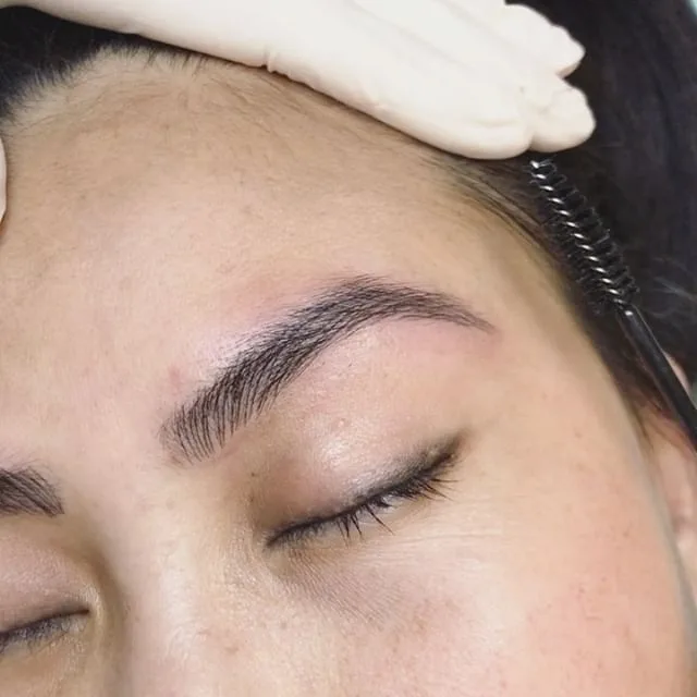 A healed feather-stroke brow being groomed with a spoolie brush held in a white-gloved practitioner's hand